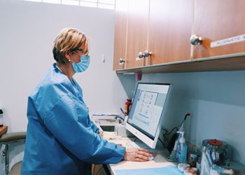 A woman in blue scrubs is standing at the computer.